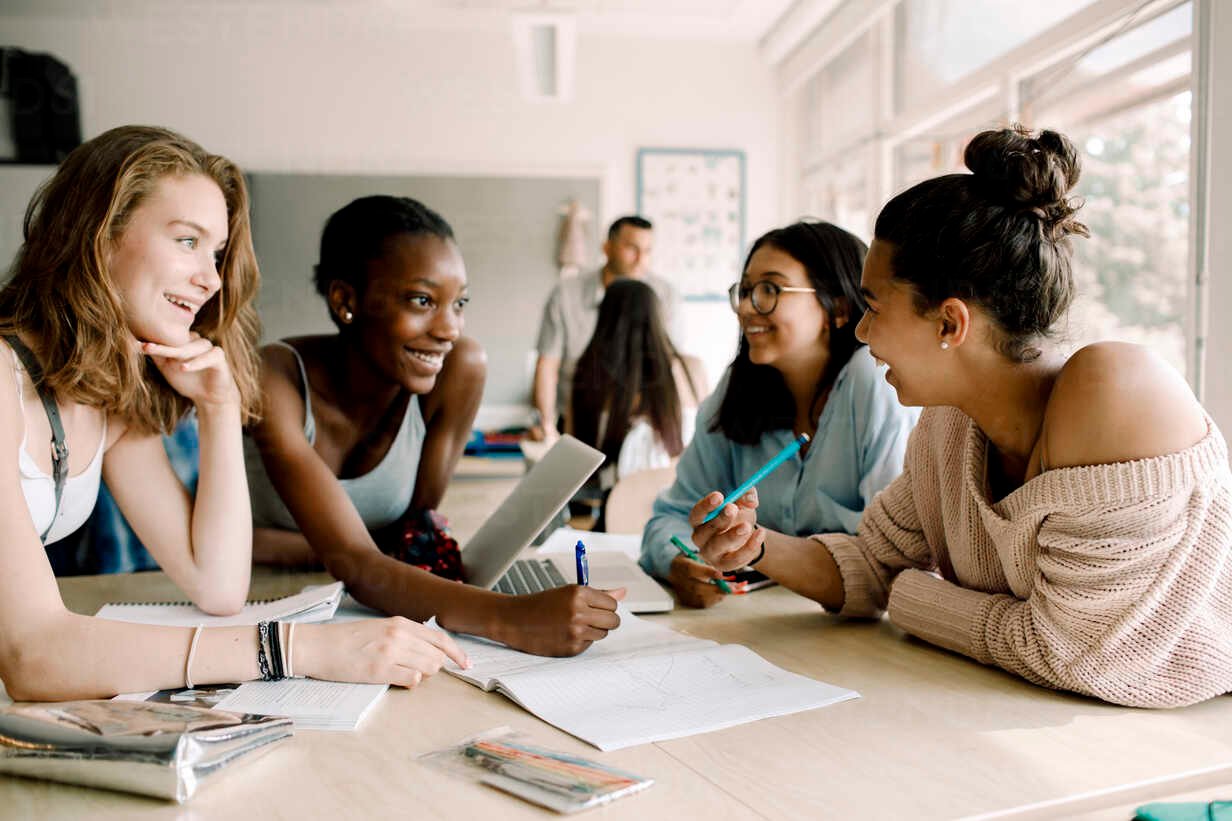 smiling-female-teenagers-discussing-while-sitting-by-table-in-classroom-MASF17378